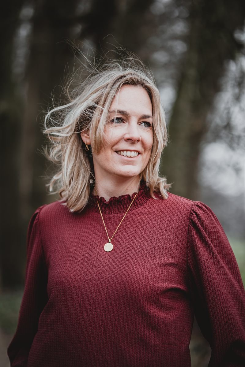 Yanieke lacht, buiten tussen de bomen, in een donkerrode trui.